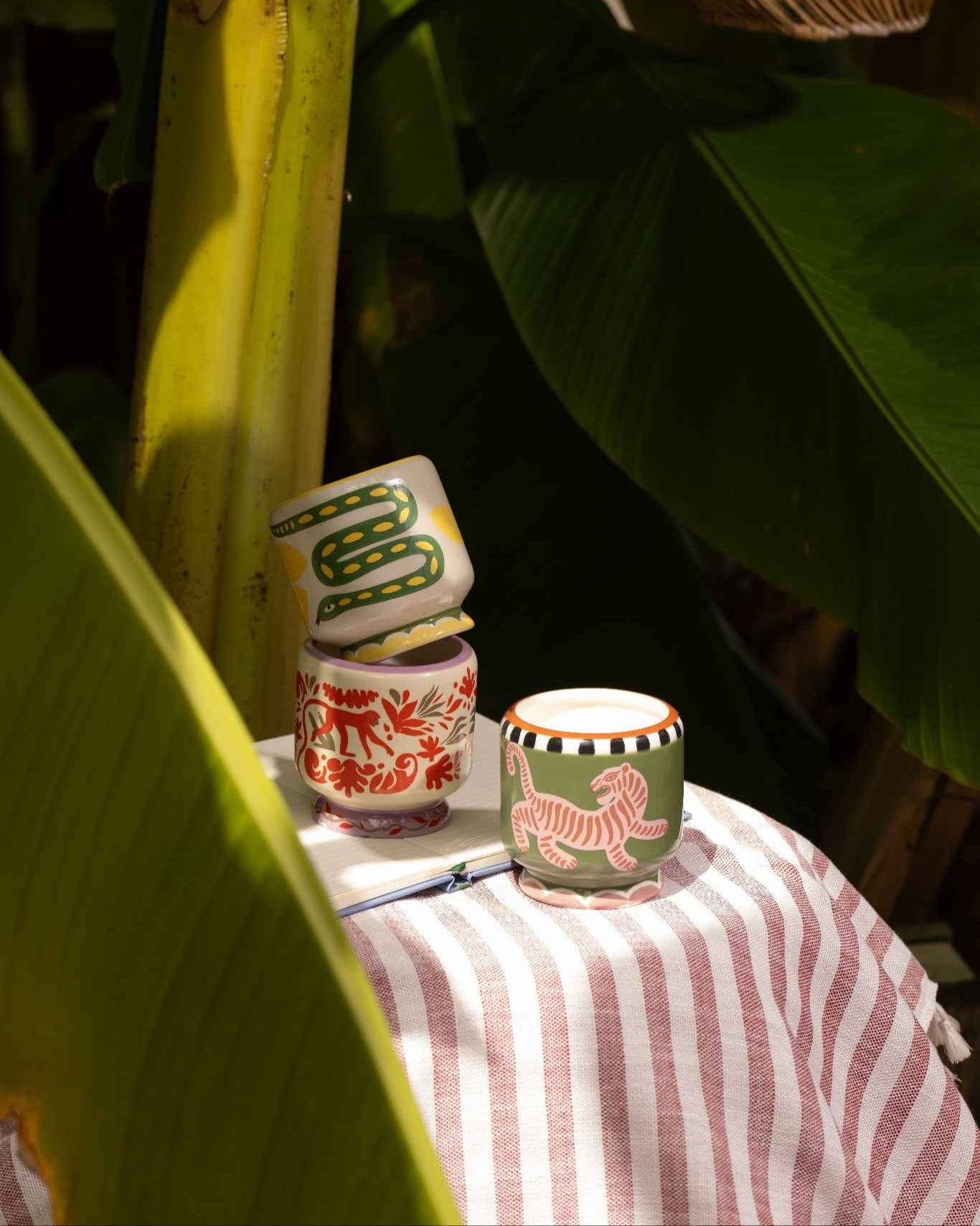 Vela en cerámica pintada a mano con motivo de tigre y detalles en negro, junto a otras tazas ilustradas.