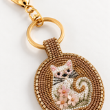 Detalle de charm con gato blanco, bigotes negros, flores rosas y bordado de abalorios dorados.