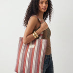 Mujer llevando una bolsa tote de jacquard con estampado de rayas en tonos rojo, granate, lila y crudo.