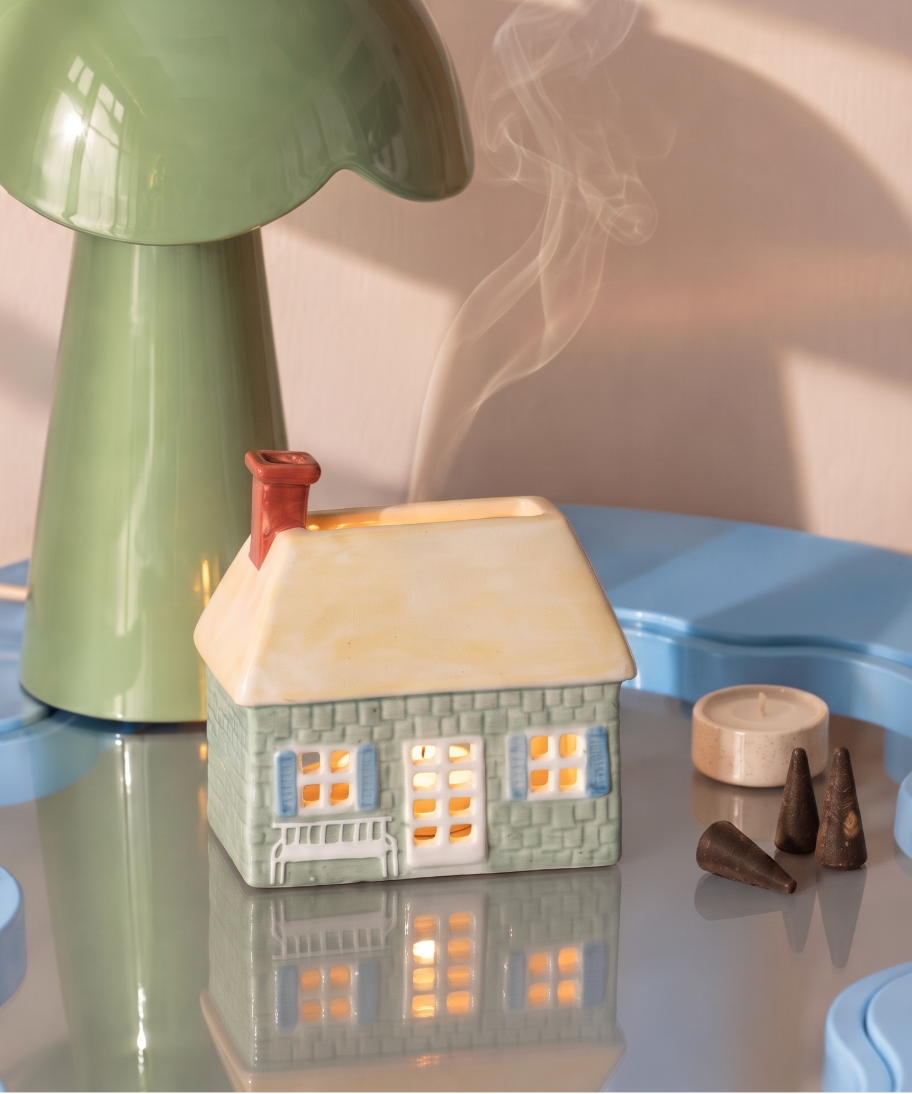 Casita de cerámica PADDYWAX verde con tejado crema, iluminada y con humo saliendo de la chimenea.