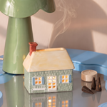 Casita de cerámica PADDYWAX verde con tejado crema, iluminada y con humo saliendo de la chimenea.