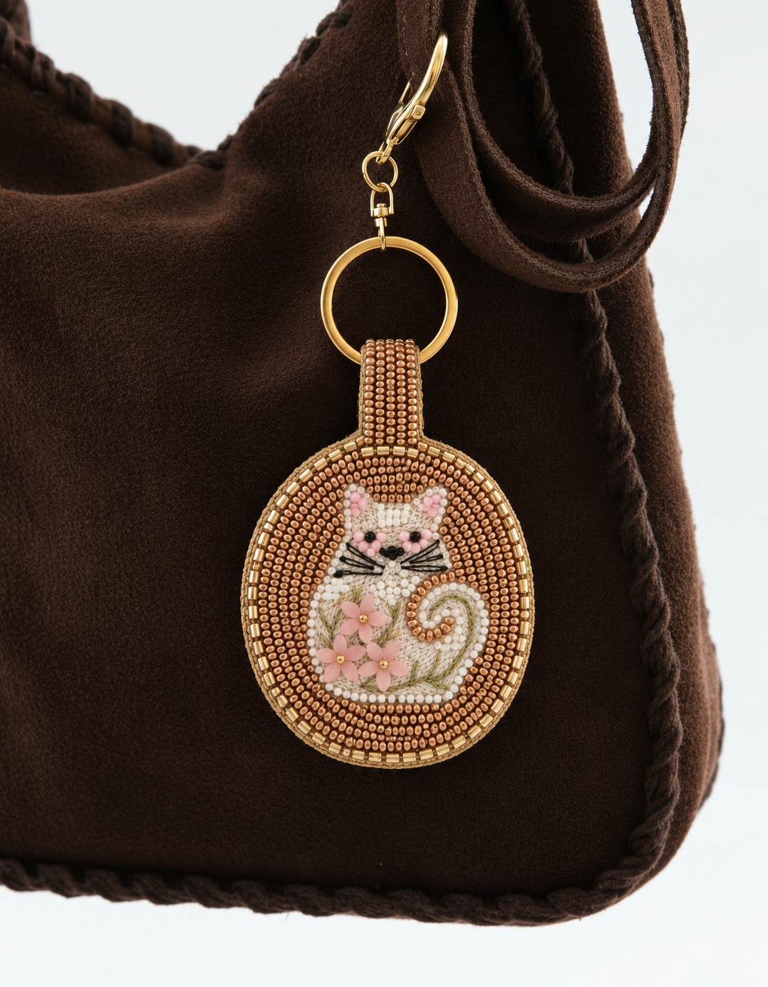 Charm circular de gato blanco con flores rosas y abalorios dorados colgado de un bolso marrón.