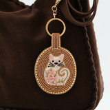 Charm circular de gato blanco con flores rosas y abalorios dorados colgado de un bolso marrón.
