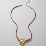 Collar de acero inoxidable con colgante de sol dorado y cordón burdeos sobre fondo blanco.