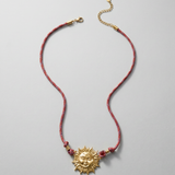 Collar de acero inoxidable con colgante de sol dorado y cordón burdeos sobre fondo blanco.
