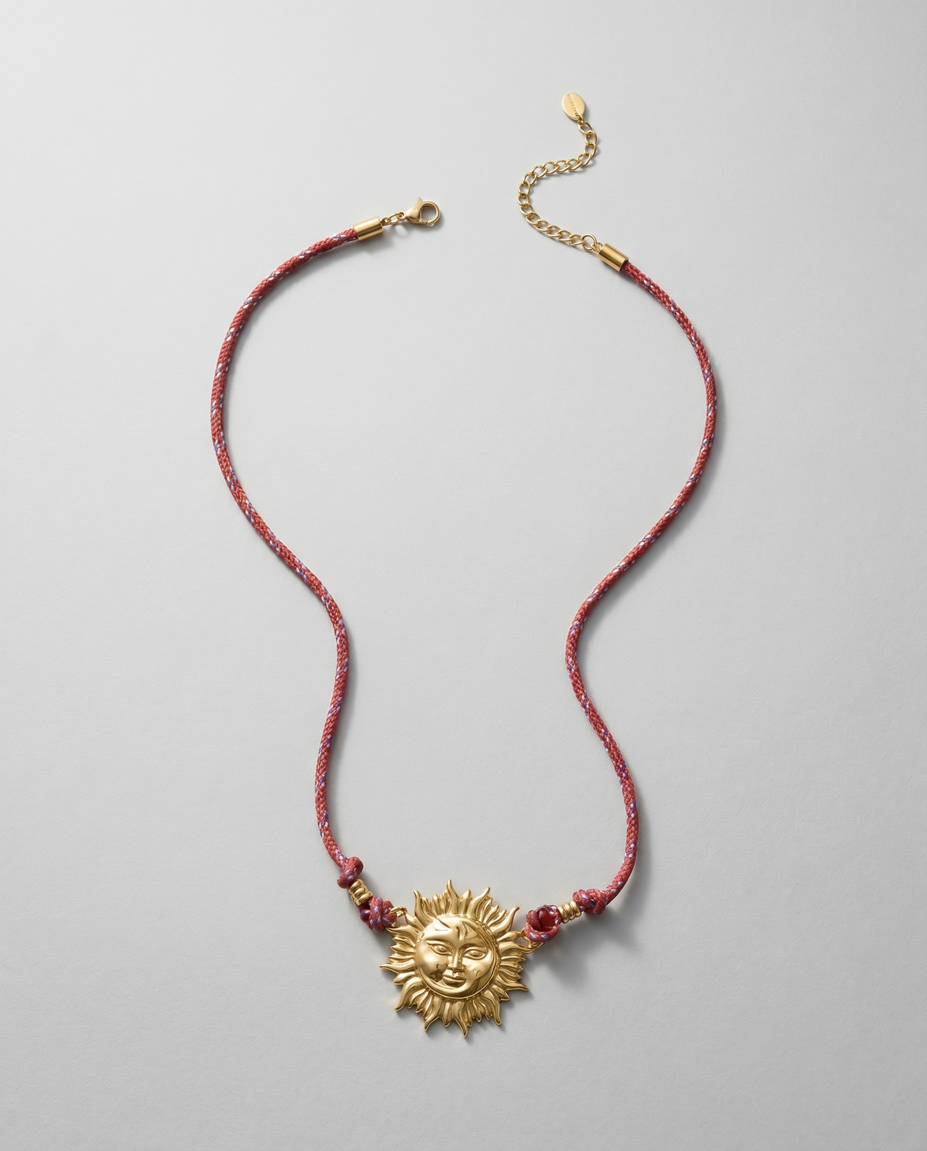 Collar de acero inoxidable con colgante de sol dorado y cordón burdeos sobre fondo blanco.