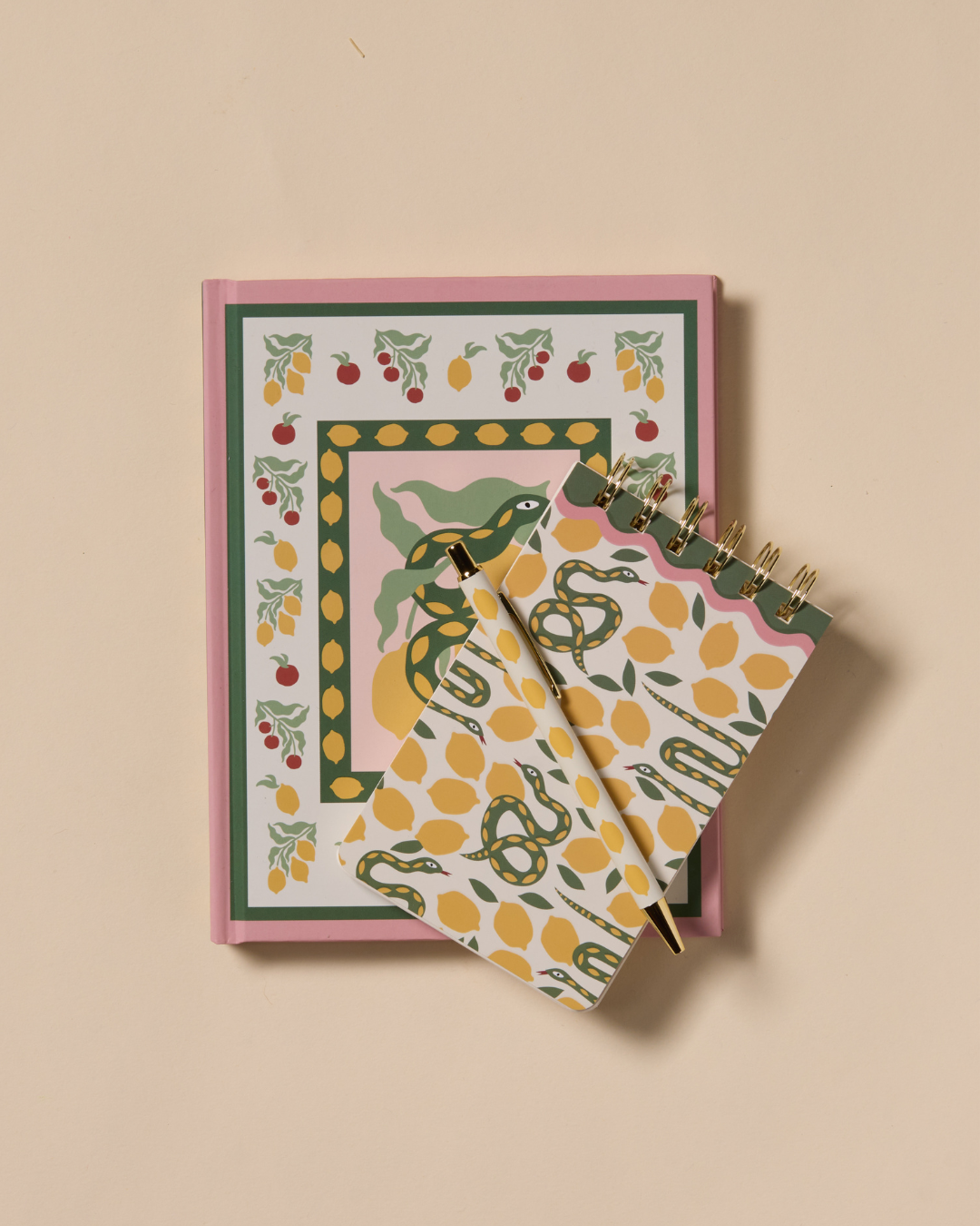 Cuaderno de notas rosa con ilustraciones de serpientes verdes y frutas, acompañado de un bloc de notas a juego con estampado de limones.
