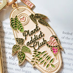 Detalle del llavero botánico con hojas verdes y rosas sobre fondo beige.