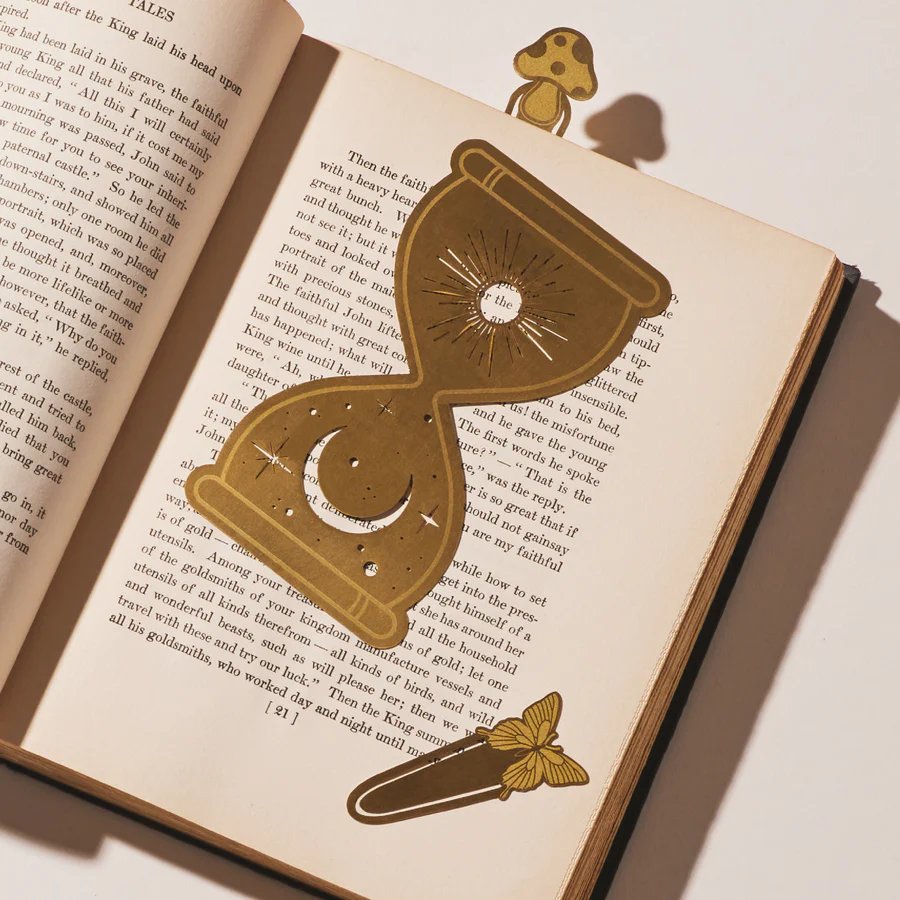 Marcapáginas de metal dorado con forma de reloj de arena sobre libro abierto, vista frontal.