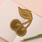Marcapáginas metálico con diseño de cerezas sobre un cuaderno abierto.