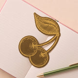 Marcapáginas metálico con diseño de cerezas sobre un cuaderno abierto.