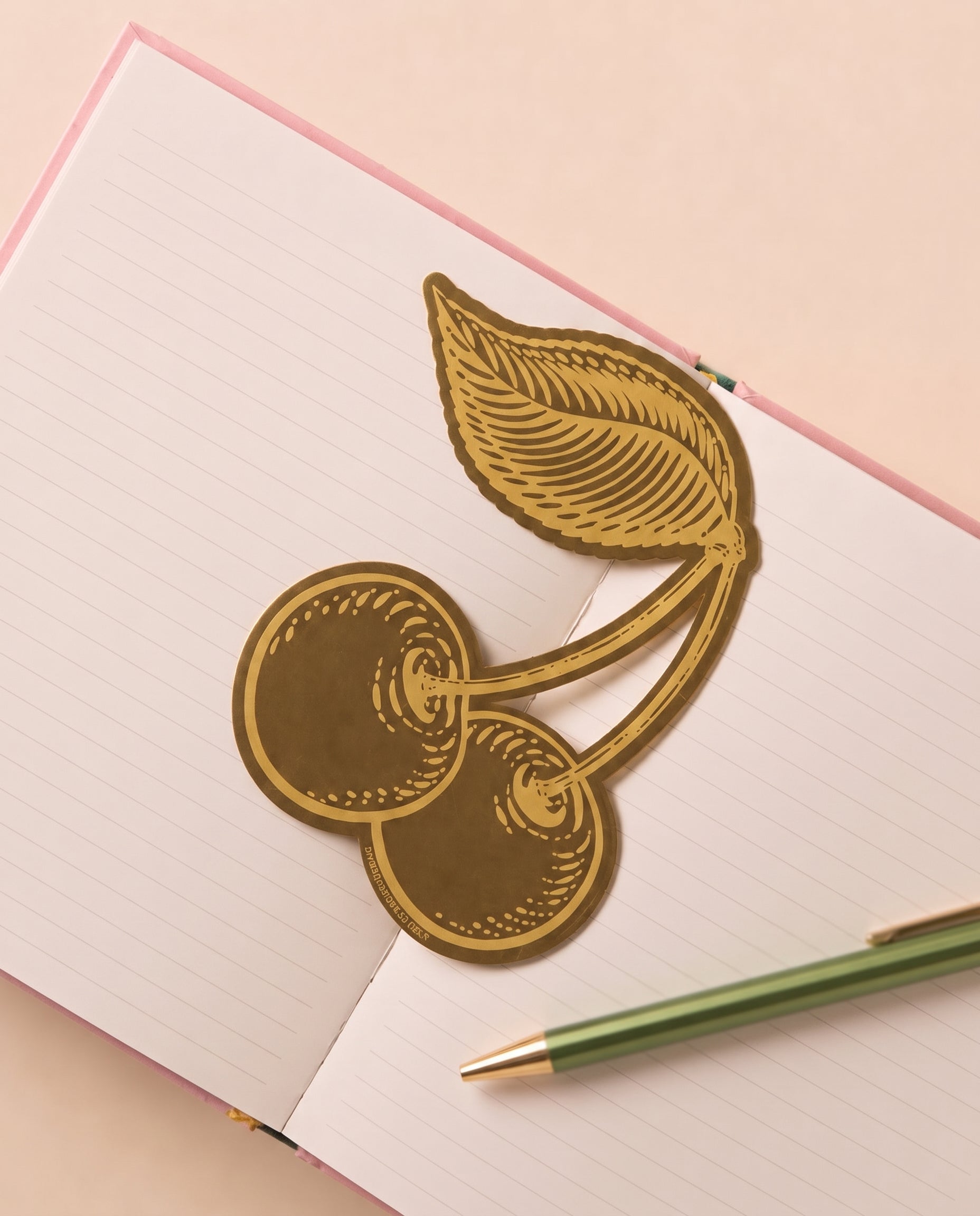Marcapáginas metálico con diseño de cerezas sobre un cuaderno abierto.