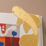 Marcapáginas metálico con forma de pez koi colocado entre las páginas de un libro.