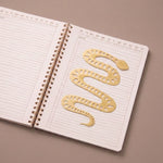 Marcapáginas metálico con forma de serpiente colocado entre las páginas de un cuaderno abierto.