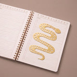 Marcapáginas metálico con forma de serpiente colocado entre las páginas de un cuaderno abierto.