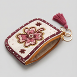 Monedero floral visto en diagonal con cremallera, lateral ocre y anilla metálica con borla granate.