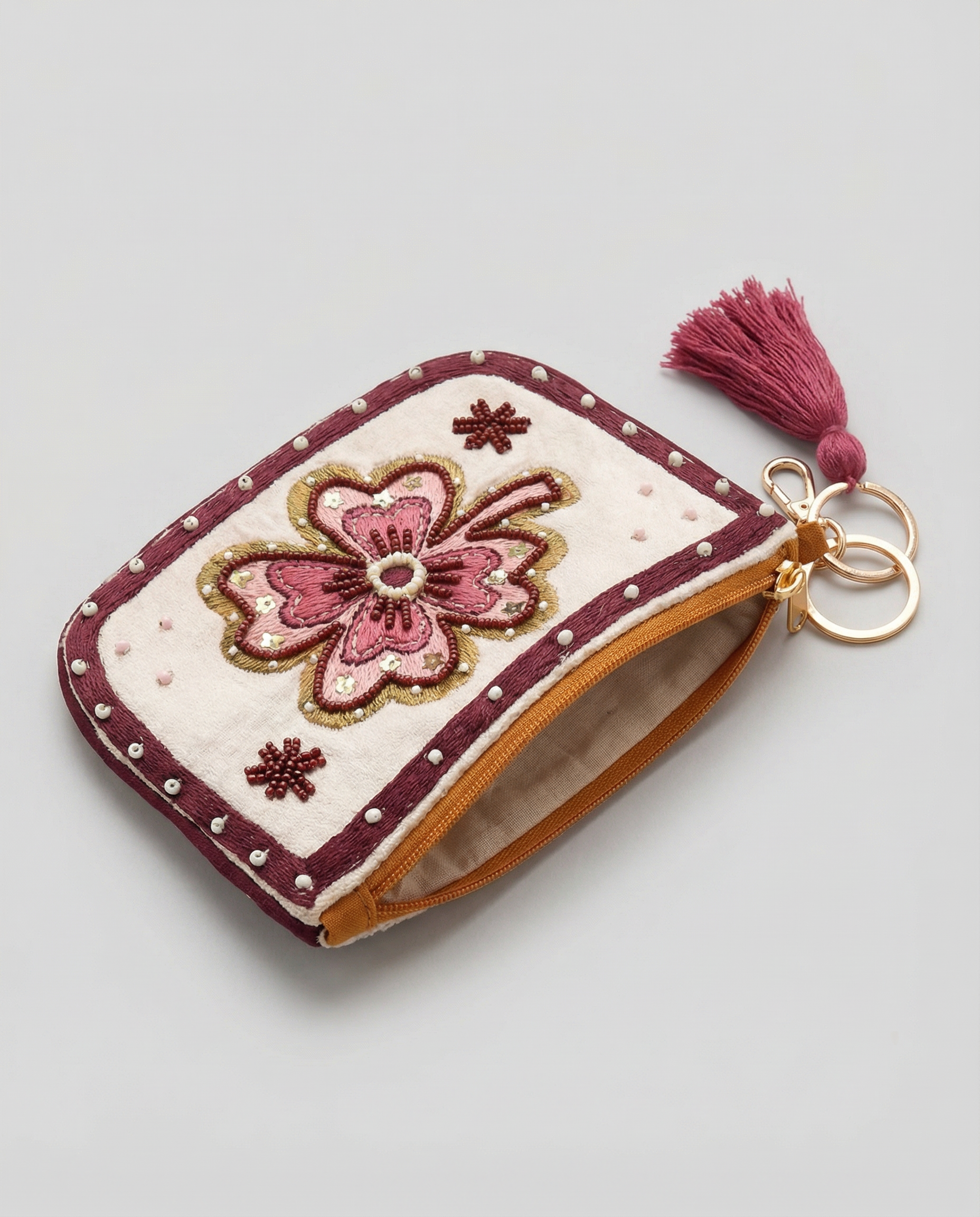 Monedero floral visto en diagonal con cremallera, lateral ocre y anilla metálica con borla granate.