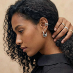 Pendientes plateados con aro y nudo texturizado en la oreja de una modelo de piel oscura y cabello rizado.