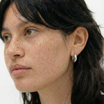 Pendientes plateados pequeños tipo aro en la oreja de una modelo con pecas, vista lateral.