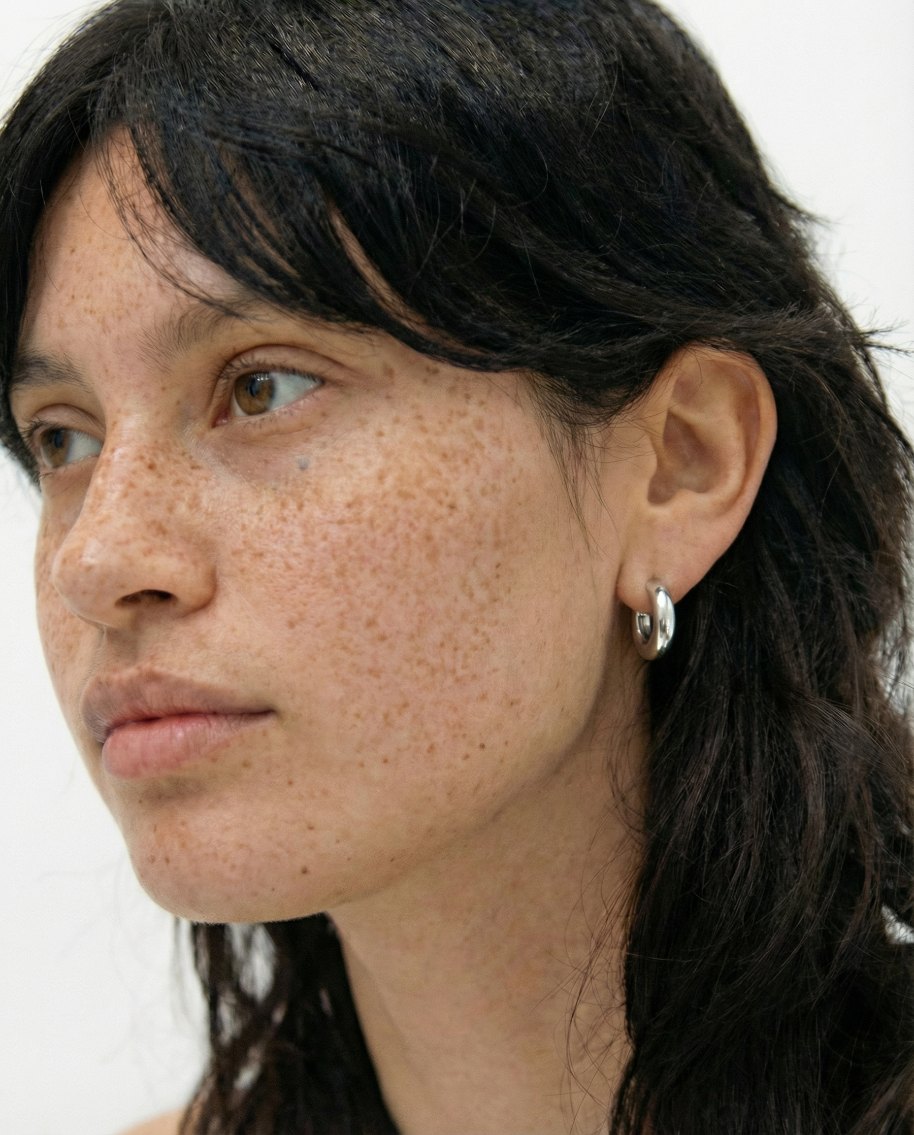 Pendientes plateados pequeños tipo aro en la oreja de una modelo con pecas, vista lateral.