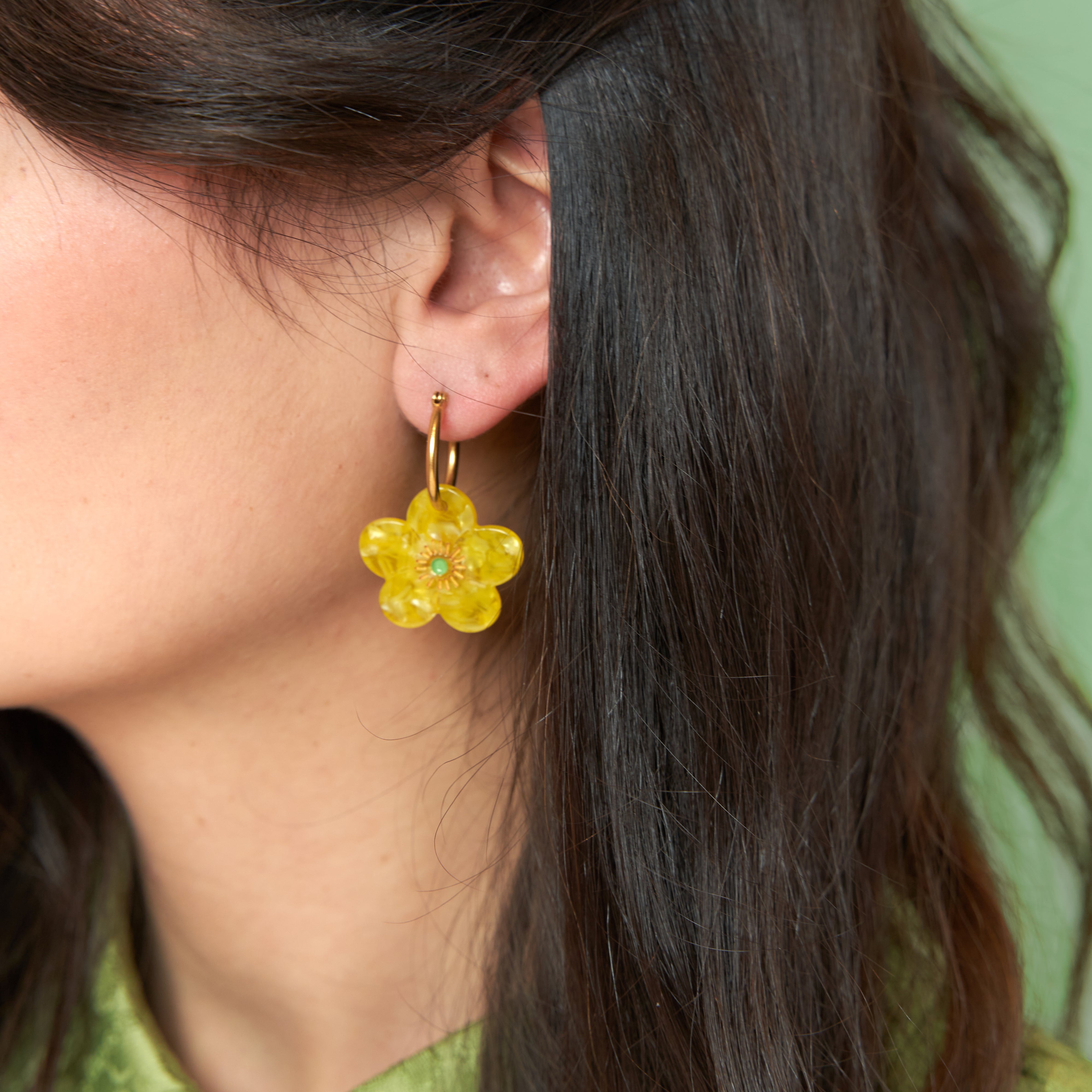 Pendiente flor Coucou Suzette bouton d'or colocado en la oreja de una modelo con fondo verde.