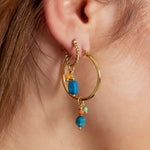Pendientes boho de aro pequeño dorado con charm colgante azul, puestos en oreja.

