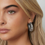 Pendiente plateado de diseño fluido y forma de aro irregular visto en la oreja de una modelo, primer plano lateral.