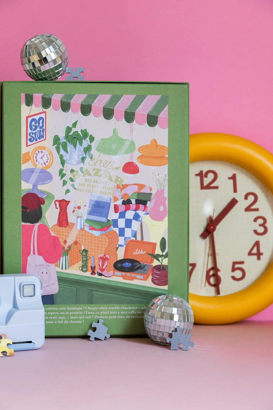 Parte trasera de la caja verde del puzzle Le Lèche-Vitrine de Jour Férié, fotografiada sobre fondo rosa con accesorios decorativos.