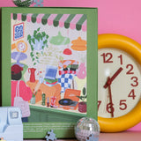 Parte trasera de la caja verde del puzzle Le Lèche-Vitrine de Jour Férié, fotografiada sobre fondo rosa con accesorios decorativos.