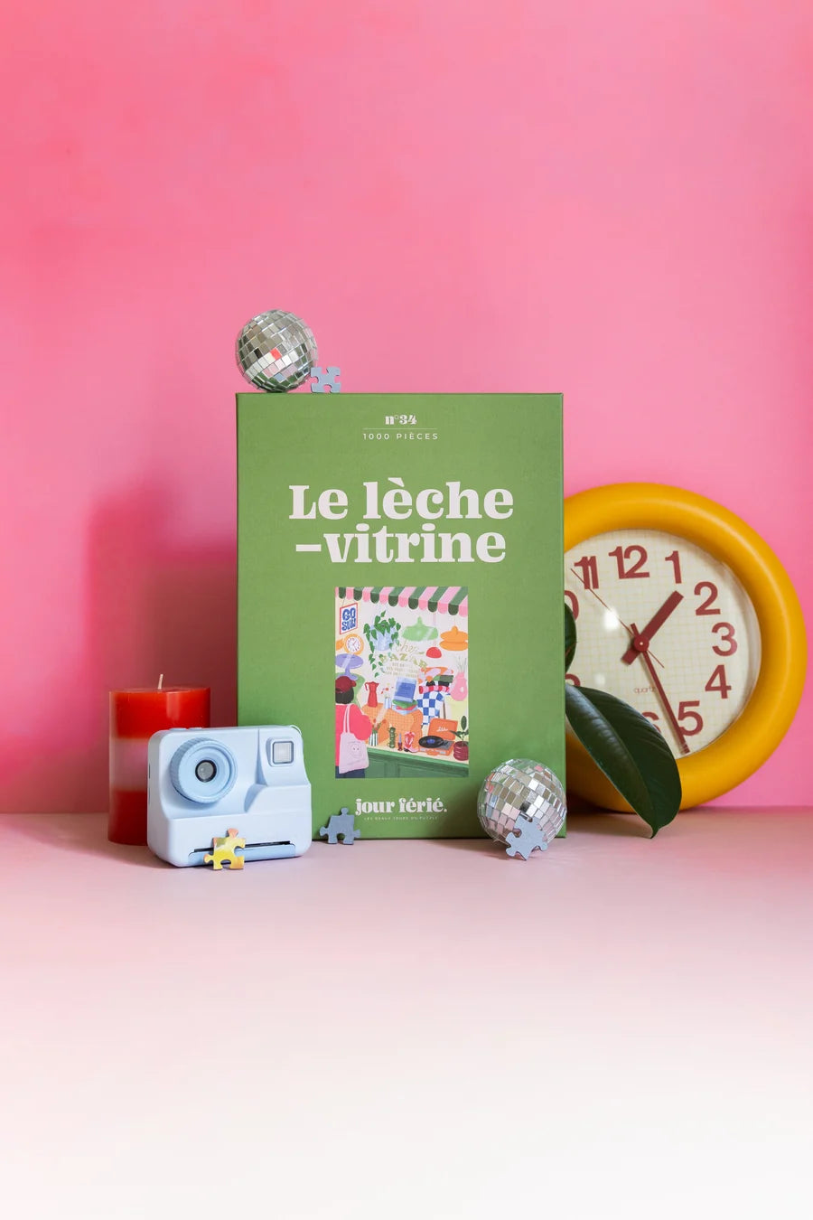 Caja verde del puzzle Le Lèche-Vitrine de 1000 piezas de Jour Férié, junto a un reloj amarillo y objetos decorativos sobre fondo rosa.