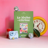 Caja verde del puzzle Le Lèche-Vitrine de 1000 piezas de Jour Férié, junto a un reloj amarillo y objetos decorativos sobre fondo rosa.