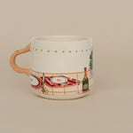Taza de cerámica ilustrada en tonos rojos y dorados con mesa navideña, vista lateral.