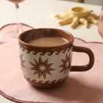 Taza marrón de cerámica con estampado de estrellas y olas sobre mantel rosa.