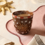 Taza marrón Sol y Estrellas sobre una mesa con mantel rosa.