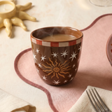 Taza marrón Sol y Estrellas sobre una mesa con mantel rosa.