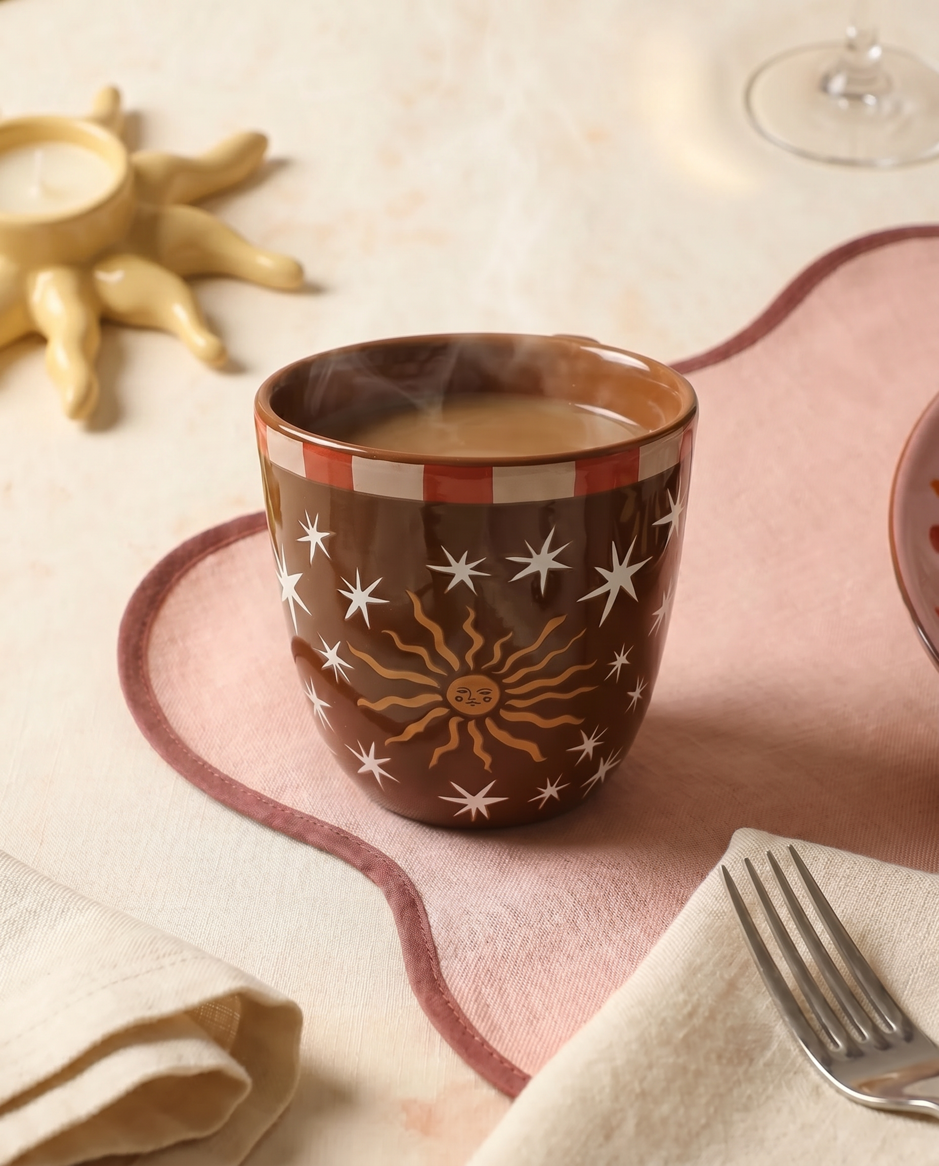 Taza marrón Sol y Estrellas sobre una mesa con mantel rosa.