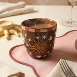 Taza de cerámica marrón con ilustración de un sol y estrellas sobre una mesa.