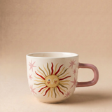 Taza de cerámica rosa con ilustración de un sol sonriente sobre fondo beige.