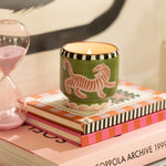 Vela aromática de soja en vaso de cerámica verde con tigre rosa, vista frontal sobre libros