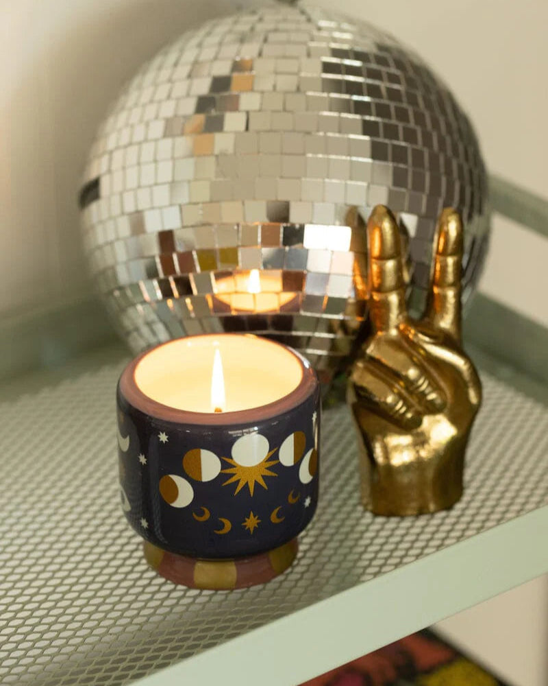 Vela de soja con dibujo de lunas y soles sobre estantería con bola de espejos y mano dorada decorativa Urban Outfitters

