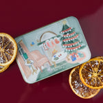 Vela aromática de Navidad PADDYWAX en lata verde salvia con ilustración de salón navideño, árbol y chimenea.