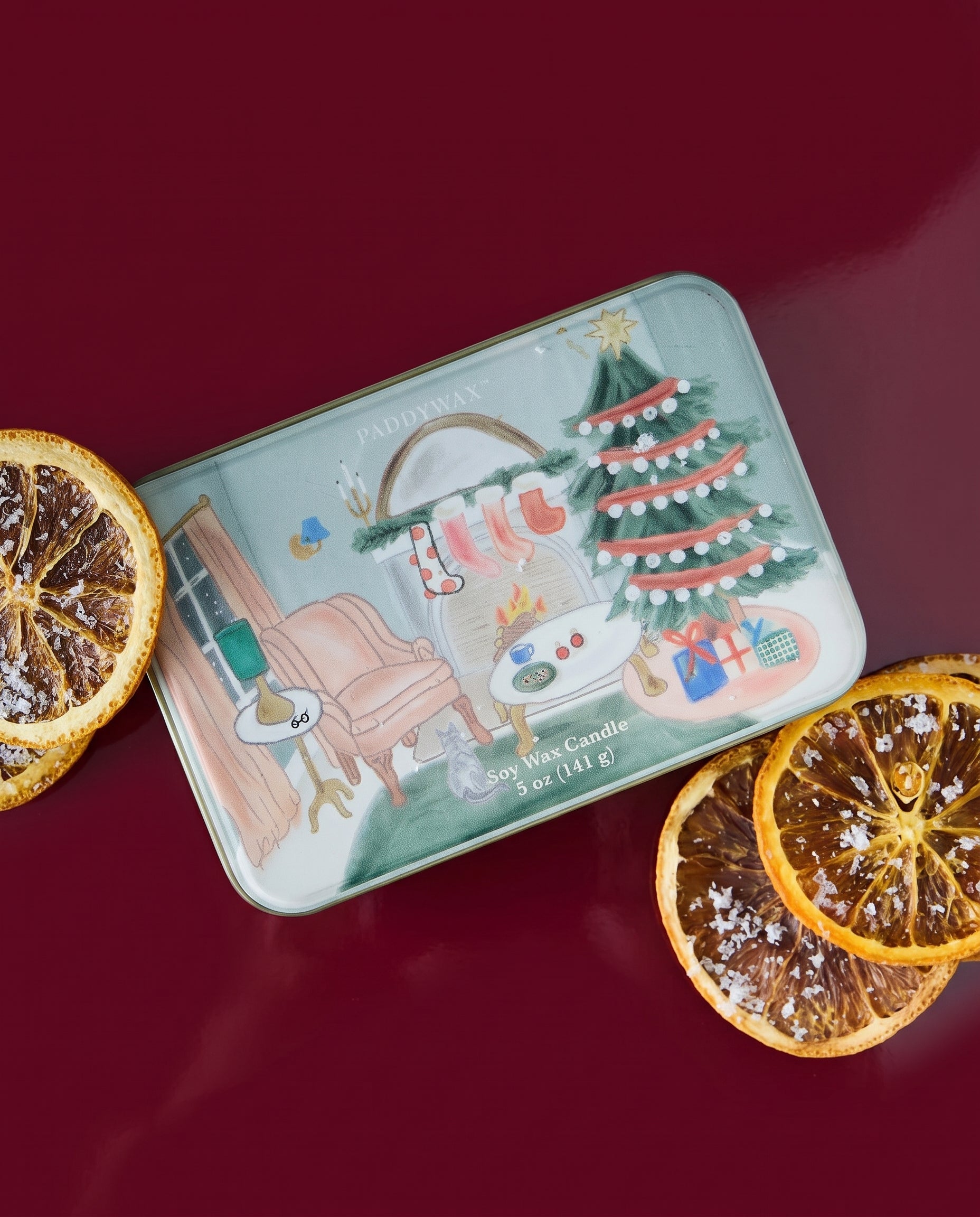 Vela aromática de Navidad PADDYWAX en lata verde salvia con ilustración de salón navideño, árbol y chimenea.