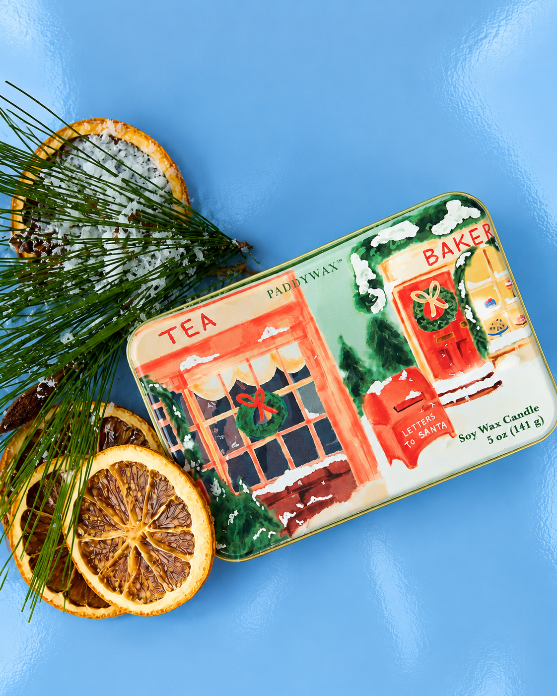 Vela aromática PADDYWAX de naranja y canela en lata metálica con ilustración de tiendas navideñas y buzón rojo.