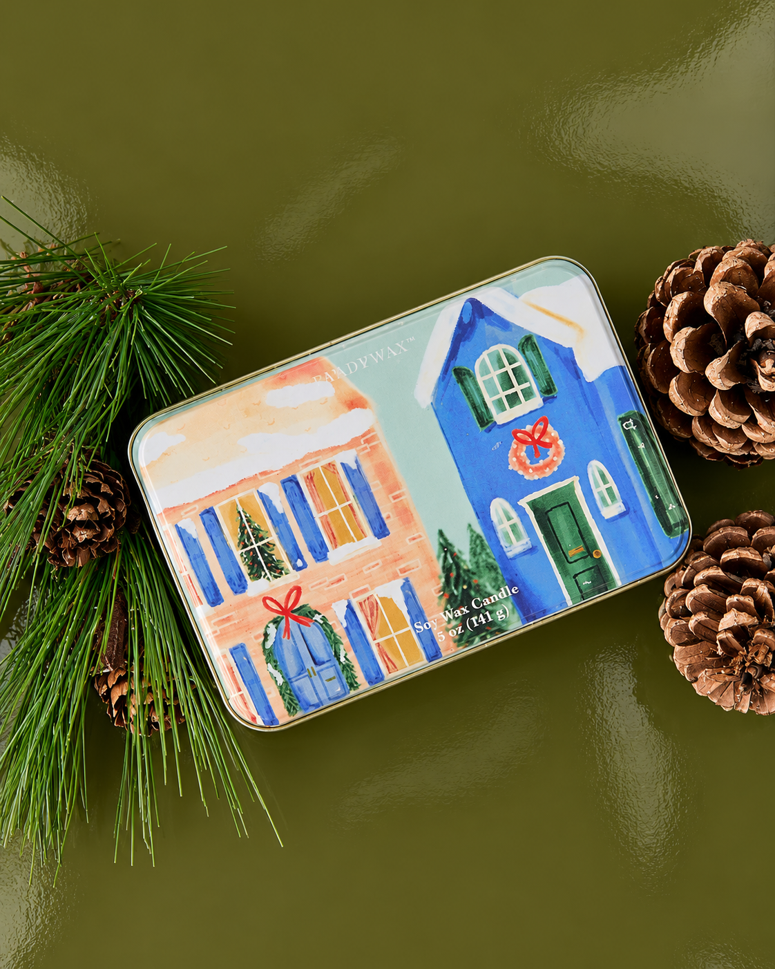 Vela aromática PADDYWAX de pino y eucalipto en lata metálica con ilustración de casas navideñas azules y rosas.