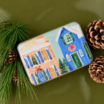 Vela aromática PADDYWAX de pino y eucalipto en lata metálica con ilustración de casas navideñas azules y rosas.