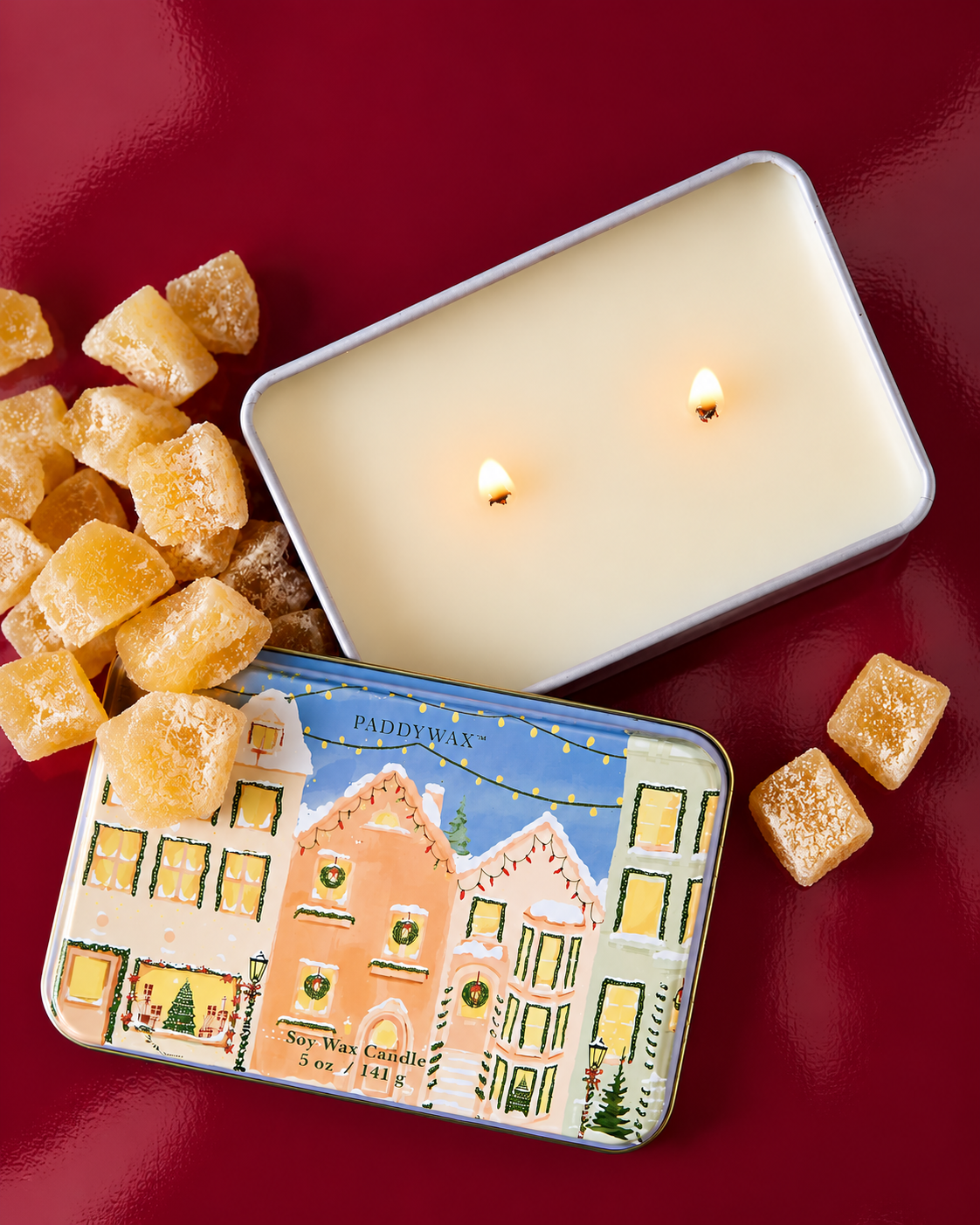 Detalle de la lata metálica PADDYWAX con casas decoradas para Navidad, sobre fondo burdeos brillante.