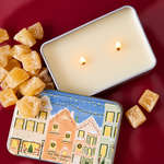 Detalle de la lata metálica PADDYWAX con casas decoradas para Navidad, sobre fondo burdeos brillante.