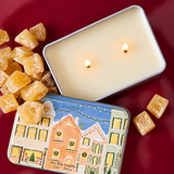 Detalle de la lata metálica PADDYWAX con casas decoradas para Navidad, sobre fondo burdeos brillante.
